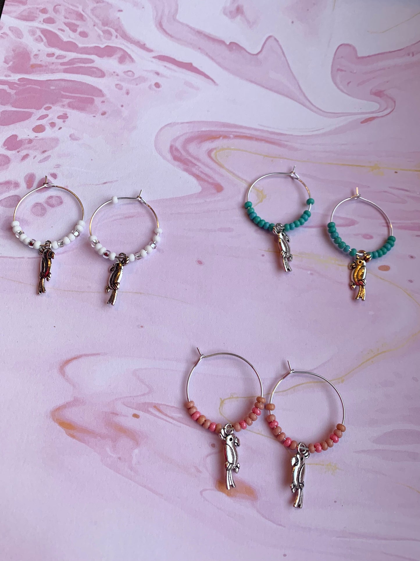 Colourful hoop earrings. Each pair has a alloy silver coloured parrot charm on silver plated hoops with different colour themed seed beads on the hoops. There is a white sparkle, turquoise and pink coral pair. Animal earrings. Bird earrings. 