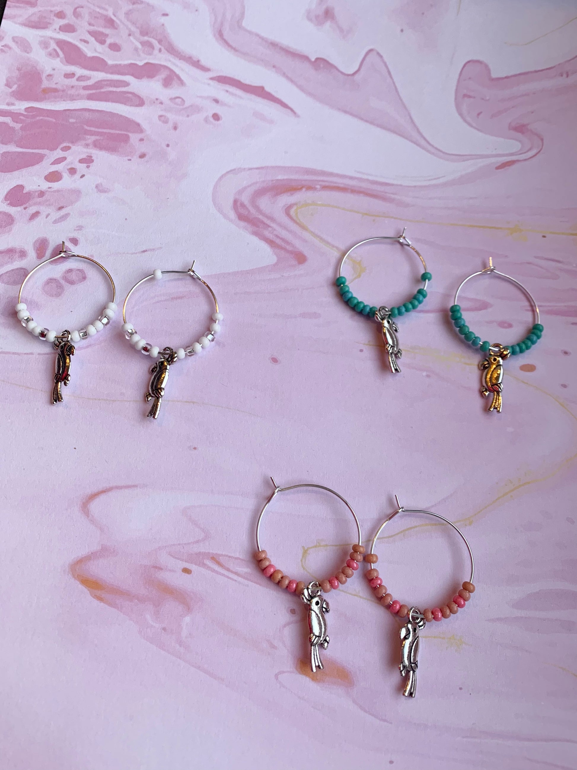Colourful hoop earrings. Each pair has a alloy silver coloured parrot charm on silver plated hoops with different colour themed seed beads on the hoops. There is a white sparkle, turquoise and pink coral pair. Animal earrings. Bird earrings. 